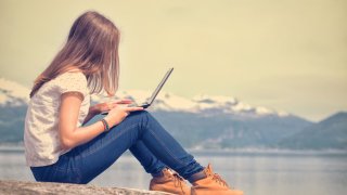 Jente sitter utendørs på en fjellknaus ved et vann, med laptop i fanget