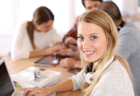En ung kvinnelig elev sitter på et bord sammen med andre elever. Hun har en PC foran seg.