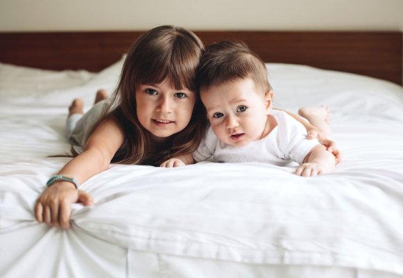 Jente og baby ligger på en seng med hvitt sengetøy, jenta holder rundt babyen.
