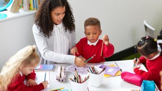 Små skolebarn og en lærer som sitter å jobber med oppgaver.