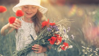 Smilende jente med hvit kjole og hvit stråhatt står i en blomstereng med en stor blomsterbukett med valmuer, prestekrager og strå
