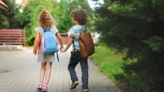 To barn, en gutt og en jente, går hånd i hånd med skolesekk på ryggen, vi ser dem bakfra