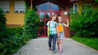 To smilende barn, en gutt og en jente, går hånd i hånd ut av en skolebygning