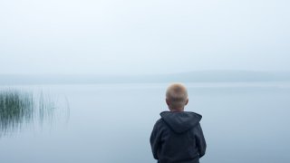 Ung gutt med blå hettejakke sitter og ser utover et disig vann, vi ser ham bakfra