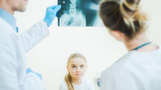 Jente titter opp på to hvitkledde personer som studerer et røntgenbilde