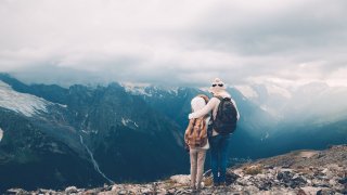 Et barn og en voksen med ryggsekk står på et fjell og ser utover fjellheimen.