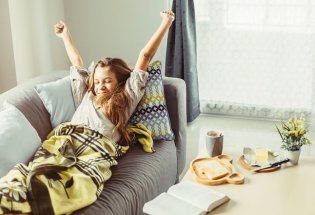 Jente som strekker seg på sofaen, hun har pledd over seg, og på bordet ved siden av står en kopp kakao, litt mat og en bok 