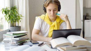 Tenåringsgutt som har hjemmeskole. Sitter med headset ved en pc med bøker rundt seg.